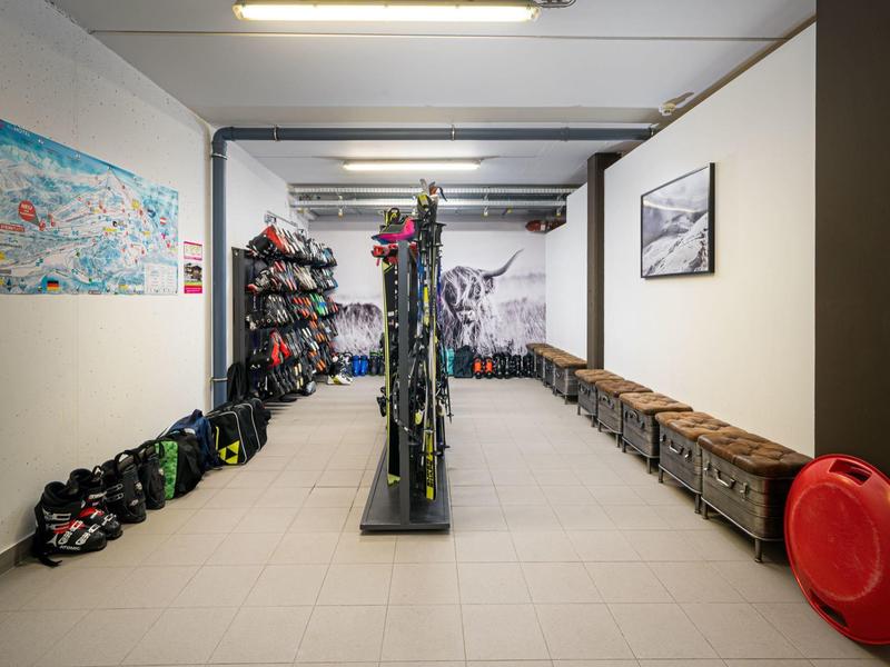 Skiraum mit Regalen, Ski, Schuhen und hölzernen Sitzbänken an weißen Wänden und grauem Boden.