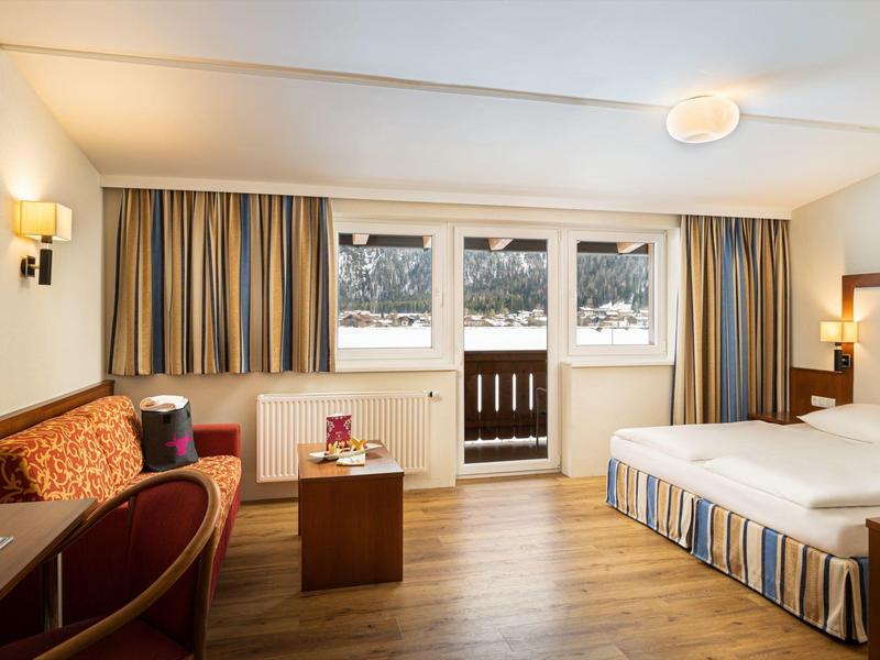 Helles Hotelzimmer mit Doppelbett, Sofa, Holzboden und großen Fenstern mit Balkonblick.