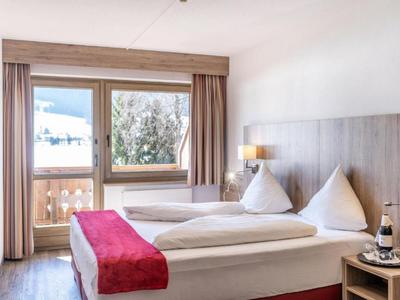 Světý hotelový pokoj s manželskou postelí, červenými doplňky a balkonem s výhledem na hory.