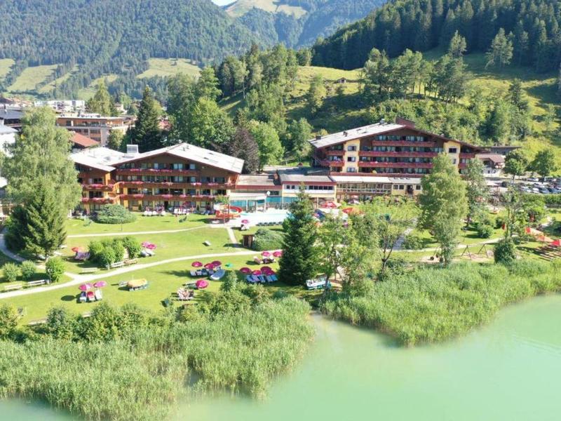 Hotelový komplex s zahradou a bazénem u břehu zeleného jezera, obklopený horami.