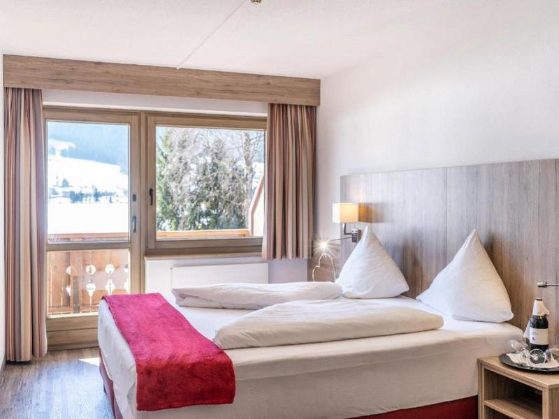 Světý hotelový pokoj s manželskou postelí, červenými doplňky a balkonem s výhledem na hory.