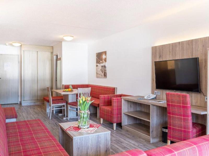 Světý hotelový pokoj s červenými kostkovanými sedacími soupravami, posezením, TV a moderním dřevěným interiérem.