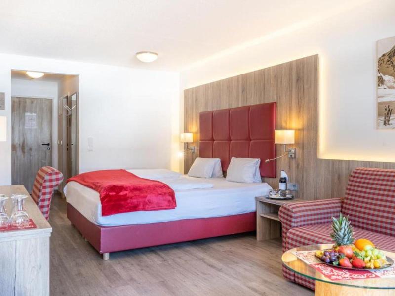 Světý hotelový pokoj s velkou postelí, červenými akcenty a posezením s ovocným talířem.