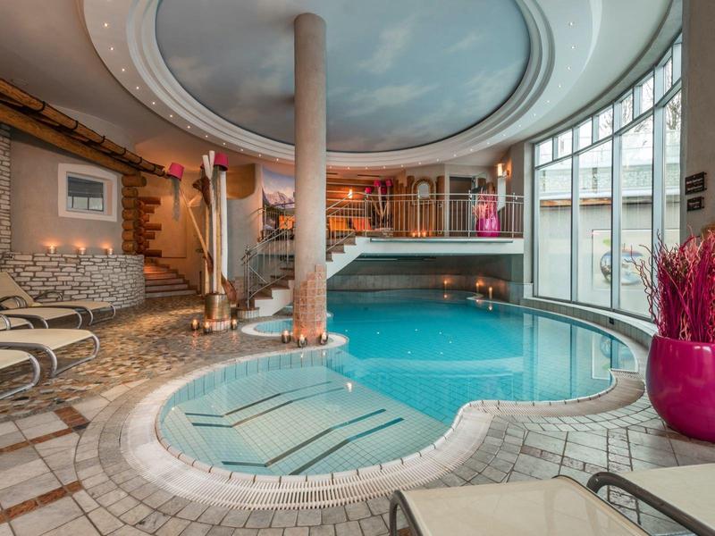 Innenraum mit rundem Schwimmbad, hellen Wänden, Fenstern und gepolsterten Liegen.