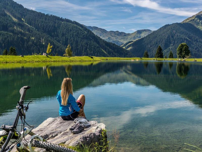 Frau sitzt auf Stein am See, umgeben von Bergen und grüner Wiese, mit Fahrrad daneben.