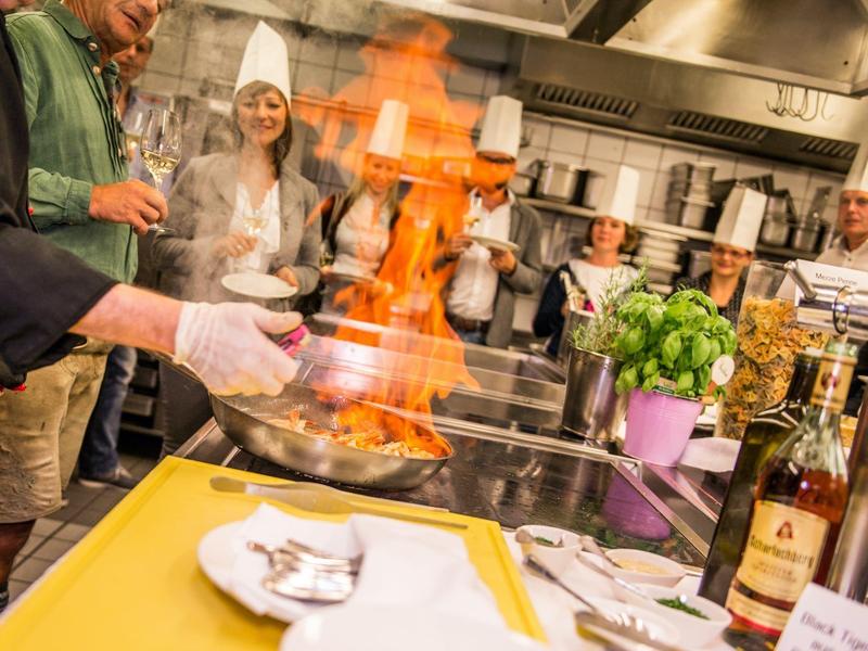 Koch zeigt flambiertes Gericht in lebhafter Restaurantküche mit lachendem Personal im Hintergrund.
