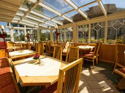 Restaurant en véranda lumineux avec tables et chaises en bois, lumière du soleil et arrangements floraux.