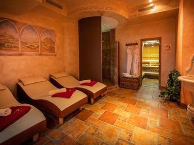 Entspannungsraum mit zwei Liegen, warmem Licht, gemusterter Fliesenboden und Wandbild mit Palmen.