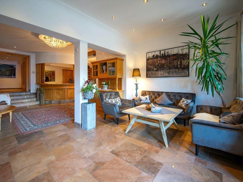 Helles Wohnzimmer mit beigen Fliesen, grauen Sofas, Holztischen und großer Pflanze neben Fenster.