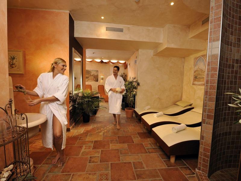 Innenansicht eines Wellnessbereichs mit Liegen, warmen Licht und zwei Personen in Bademänteln.