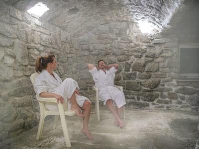 Zwei Frauen in weißen Bademänteln sitzen entspannt in einem steinernen Raum mit gedämpftem Licht.