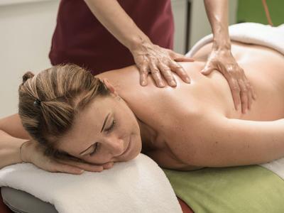 Frau entspannt auf Massagetisch, bekommt Rückenmassage von Person mit rotbraunem Oberteil.