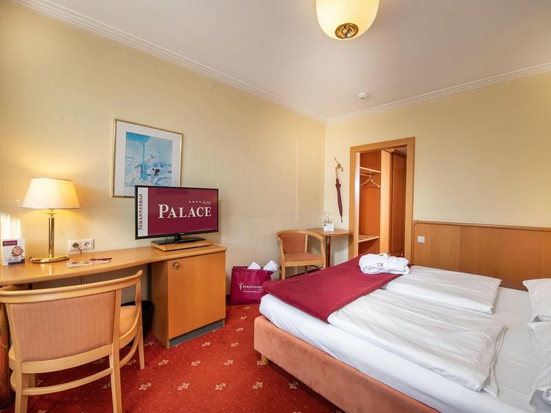 Gemütliches Hotelzimmer mit Doppelbett, Schreibtisch, Fernseher und warmem Licht, rote Teppichfarbe.