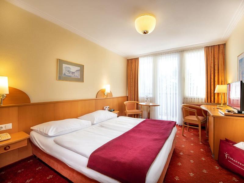 Hell eingerichtetes Hotelzimmer mit Doppelbett, roten Akzenten und Balkonfenster.