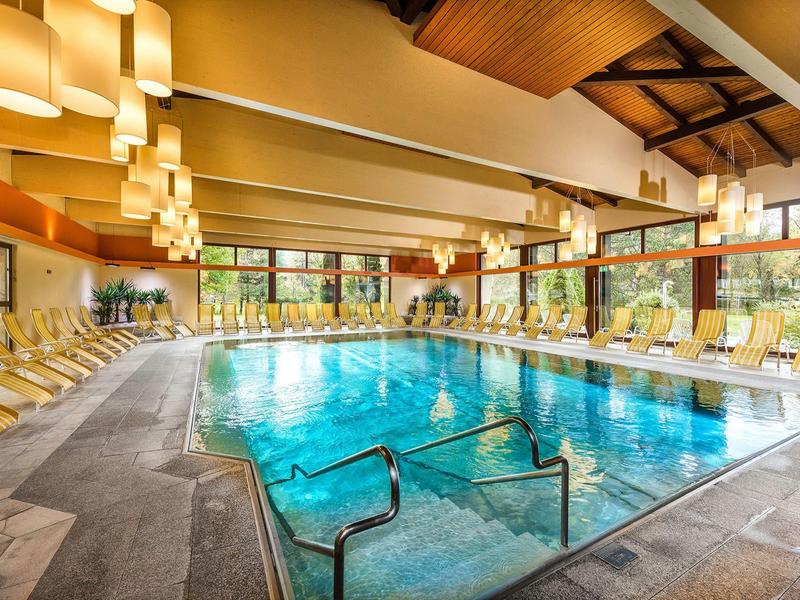 Großer Hallenbad mit Wasserbecken, Holzdecke, vielen Liegestühlen und großen Fenstern.