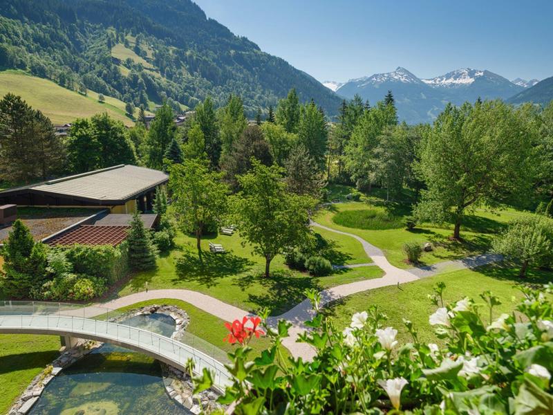 Grüner Garten mit Wegen, Blumen und Bäumen vor bewaldeten Bergen und Schneebergen im Hintergrund.