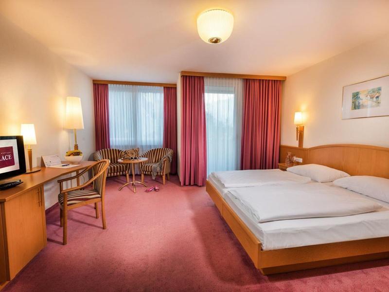 Gemütliches Hotelzimmer mit Doppelbett, Schreibtisch, Stühlen, Teppichboden und Fenster mit roten Vorhängen.