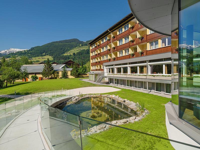 Moderner Hotelbau mit Glasfront und umlaufendem Balkon, grüner Wiese, Bergen im Hintergrund unter blauem Himmel.
