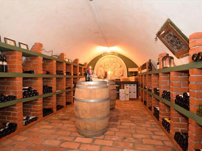 Weinkeller mit Regalen voller Weinflaschen, Tonboden und großem Holzfass in der Mitte.