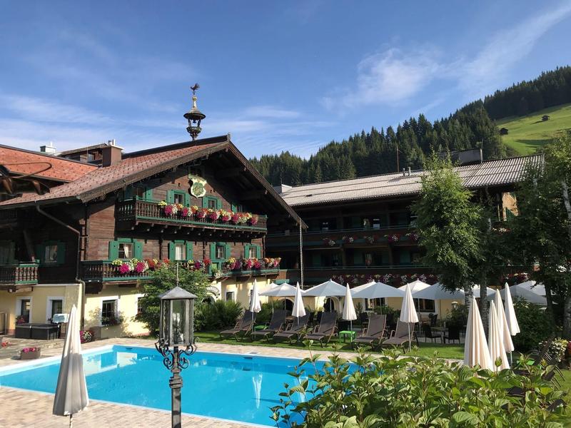 Bauernhaus im Alpenstil mit Blumenkästen, Pool und weißen Sonnenschirmen vor bewaldeten Hügeln.