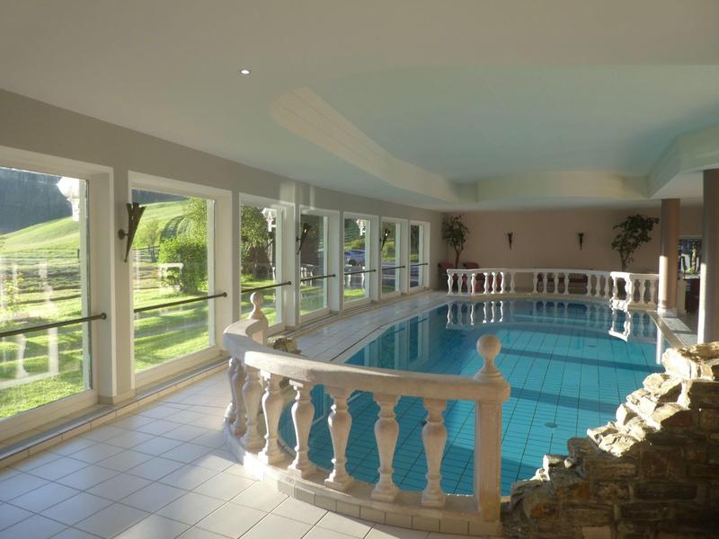Innenraum mit großem Schwimmbecken, breiten Fenstern und weißer Balustrade, hell erleuchtet.