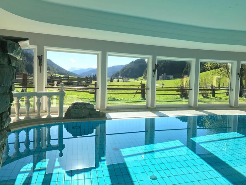 Hell beleuchteter Innenpool mit großen Fenstern und Blick auf grüne Berge und Wiesen.