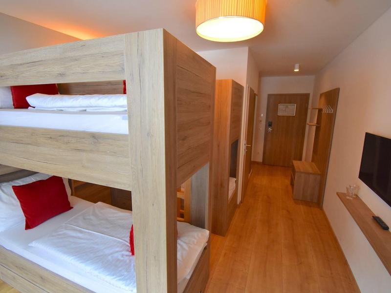 Chambre d'hôtel avec lits superposés en bois clair, coussins rouges et télévision écran plat au mur.