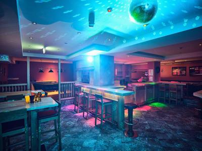 Leere Bar mit Holztischen, farbiger Beleuchtung und Spiegelkugel an der Decke in dunklem Raum.