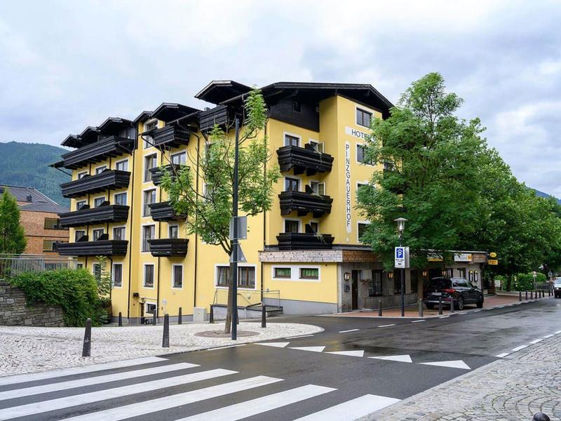 Gelbes mehrstöckiges Gebäude mit Balkonen an einer Kreuzung, umgeben von Bäumen und Bergen.