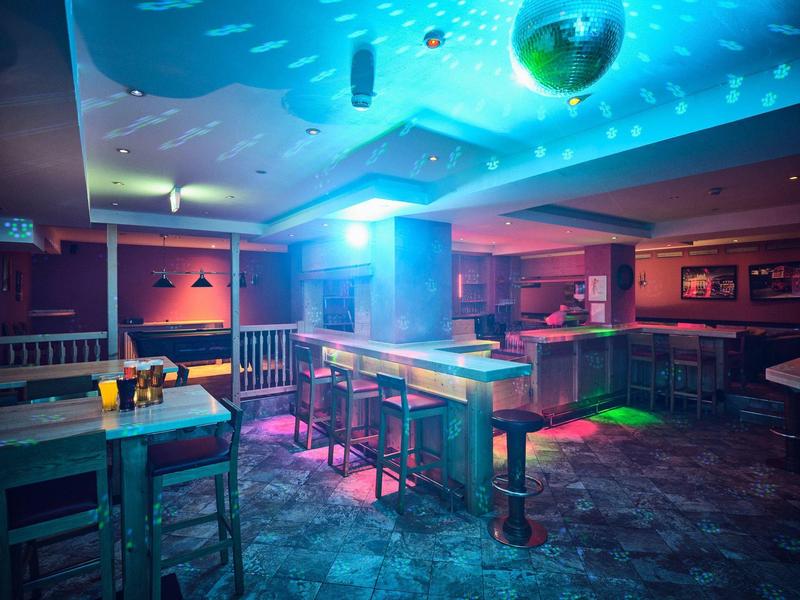 Leere Bar mit Holztischen, farbiger Beleuchtung und Spiegelkugel an der Decke in dunklem Raum.