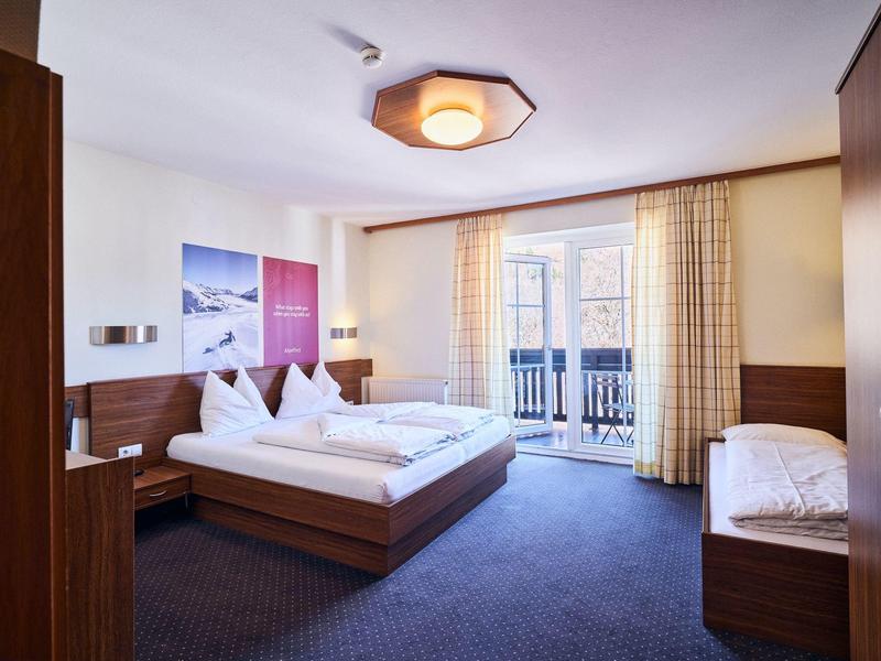 Helles Schlafzimmer mit Doppelbett, Einzelbett, blauem Teppich und Balkon durch große Fenster.