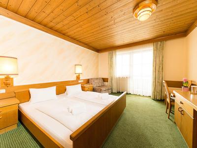 Helles Hotelzimmer mit Holzdecke, Doppelbett, grünem Teppich und Fenster mit Gardinen.
