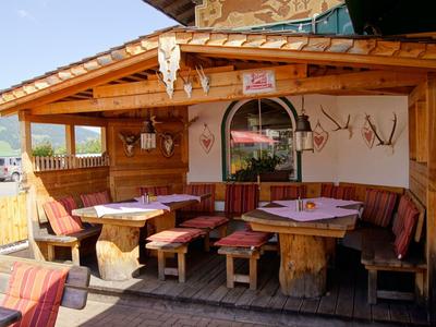 Rustikale Terrasse mit Holzmöbeln und rotem Polster vor einem Hotel bei Bergkulisse.