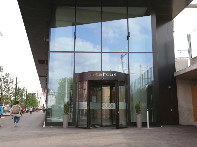 Wejście do nowoczesnego hotelu z dużymi szklanymi fasadami i okrągłymi drzwiami automatycznymi.