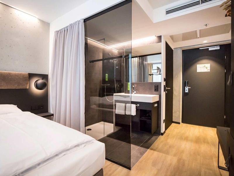 Nowoczesny pokój hotelowy z podwójnym łóżkiem, szklaną kabiną prysznicową i minimalistycznym wystrojem.