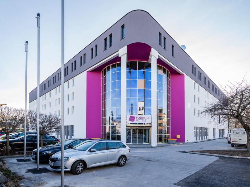 Bâtiment moderne de trois étages avec façade en verre et accents roses, parkings devant.