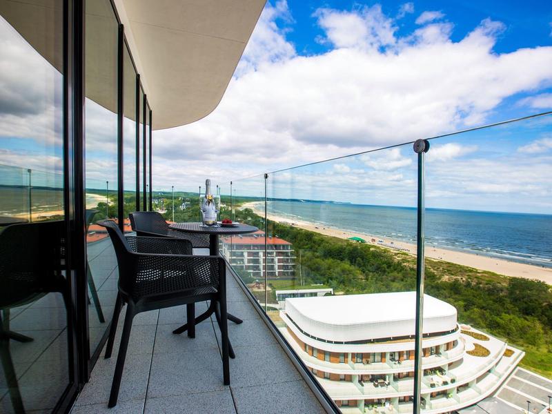 Balkon mit Glasgeländer, Stühlen und Meerblick an einem sonnigen Tag mit Wolken.
