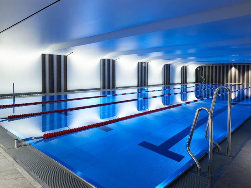 Modernes Hallenbad mit beleuchtetem Becken, Bahnmarkierungen und Edelstahlleiter unter grauem Himmel.