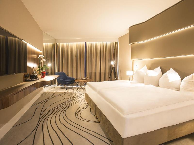 Modernes Hotelzimmer mit großem Bett, Teppich mit Muster und Vorhängen an der Fensterfront.