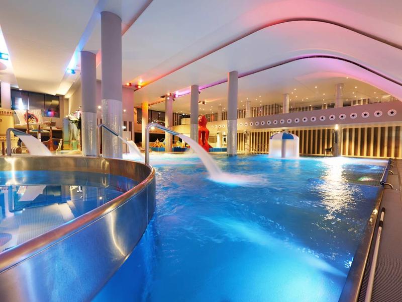 Innenbad mit großem Pool, Wasserfallspeiern, glatter Metallrutsche und warmem Licht.