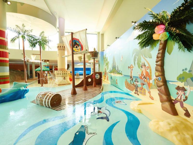 Innenraum mit Piratenschiff-Spielplatz, bunten Wellenmustern und künstlichen Palmen, helle Farben.