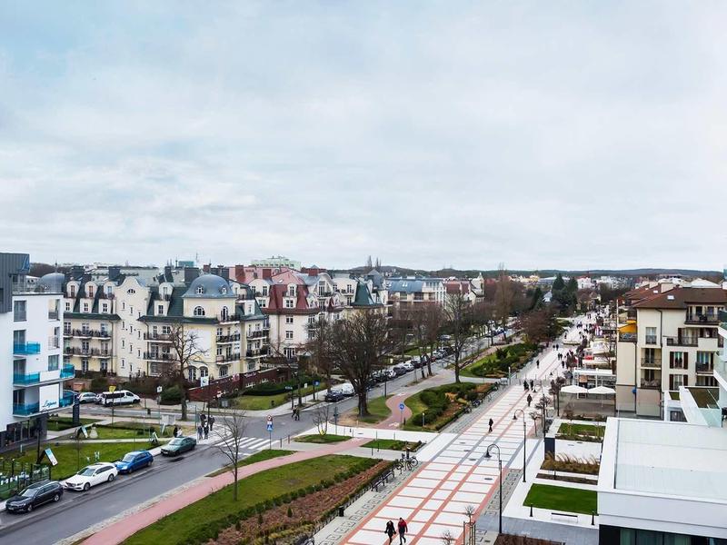 Blick auf eine moderne Stadtstraße mit Gebäuden, breiten Gehwegen und einzelnen Bäumen.