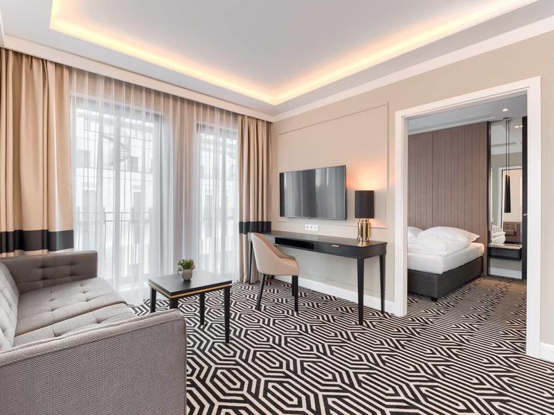 Helles Wohnzimmer mit grauem Sofa, großem Fenster, geometrischem Teppich und Fernseher an der Wand.