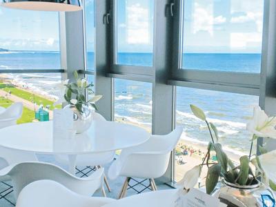 Heller Essbereich mit weißen Stühlen und runder Tisch vor großen Fenstern mit Meerblick.