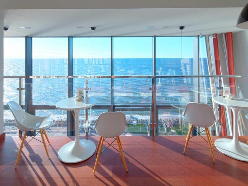 Moderner Raum mit weißen Tischen, Stühlen und großem Fenster mit Meerblick bei Tageslicht.