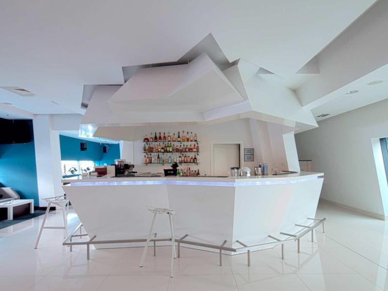 Moderner weißer Barbereich mit geometrischer Decke und transparenten Barhockern in hellem Raum.