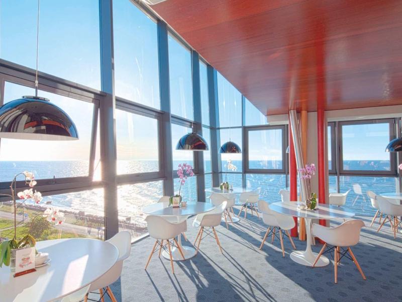 Helles Café mit großen Fenstern, weißen Tischen, runden Stühlen und Blick aufs Meer.