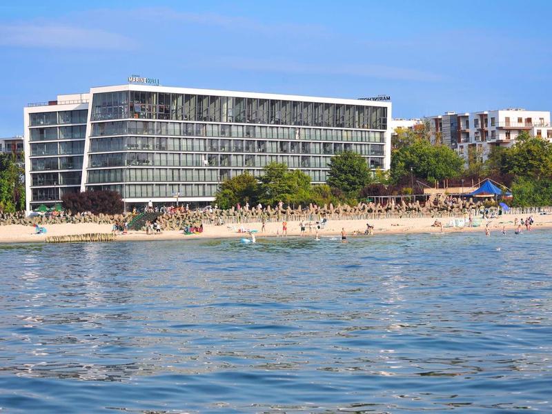 Modernes Hotelgebäude am Strand mit Sand, Palmen und klarem blauem Meer im Vordergrund.