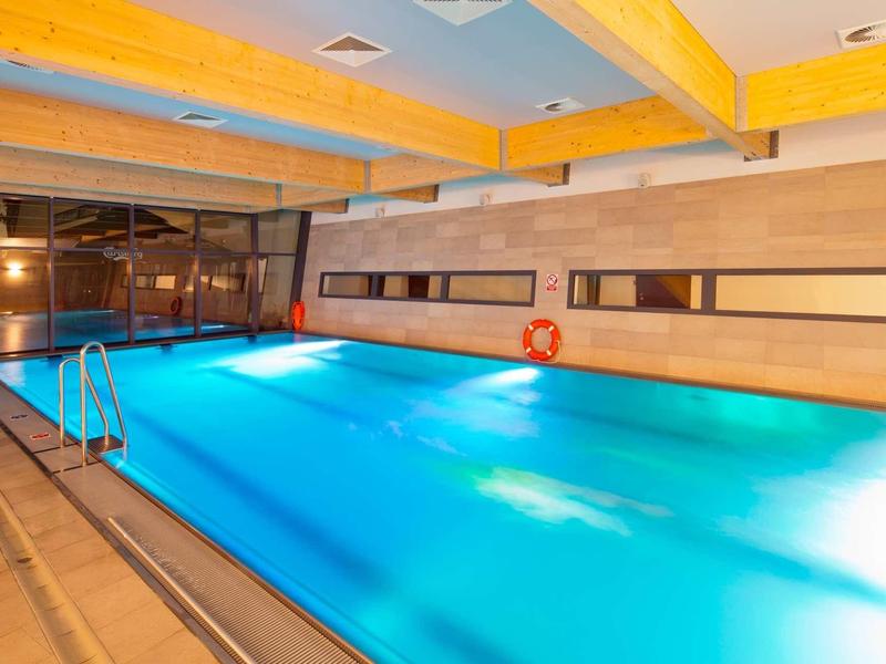 Helles Hallenbad mit blauem Wasser, Holzdeckenbalken und großen Fenstern an der rechten Wand.