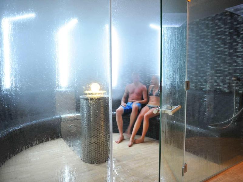 Innenraum einer feuchten Sauna mit zwei sitzenden Personen hinter Glas und hellen Leuchten.